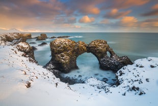 Snow covers the rugged sea arch of Gatklettur in Arnarstapi, Iceland at sunset.