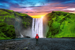 Traveler in red jacket stands in front of Skogafoss Waterfall, framed by cliffs and a colorful sunset in South Iceland.